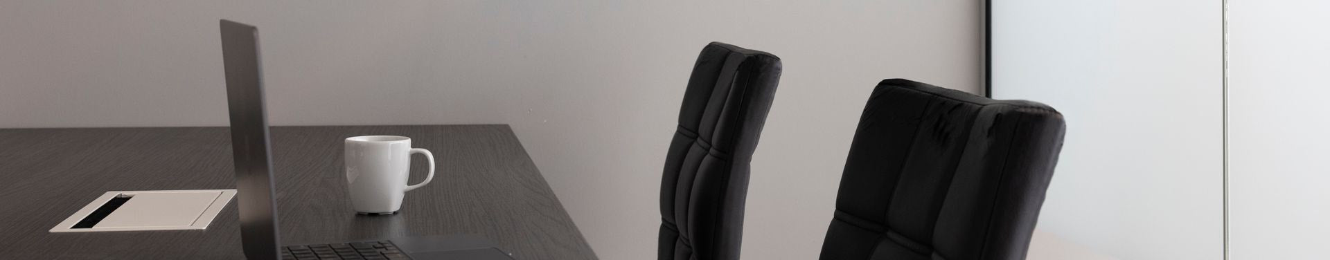 A modern office meeting setup with a dark conference table, two black leather chairs, a white coffee mug, and a laptop on a light gray background.