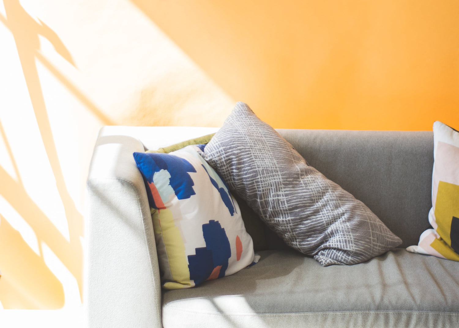 A gray fabric sofa in a brightly lit room with a warm orange wall, featuring three decorative throw pillows including a blue, yellow, and white patterned pillow, a gray textured pillow, and part of a yellow-and-white pillow.